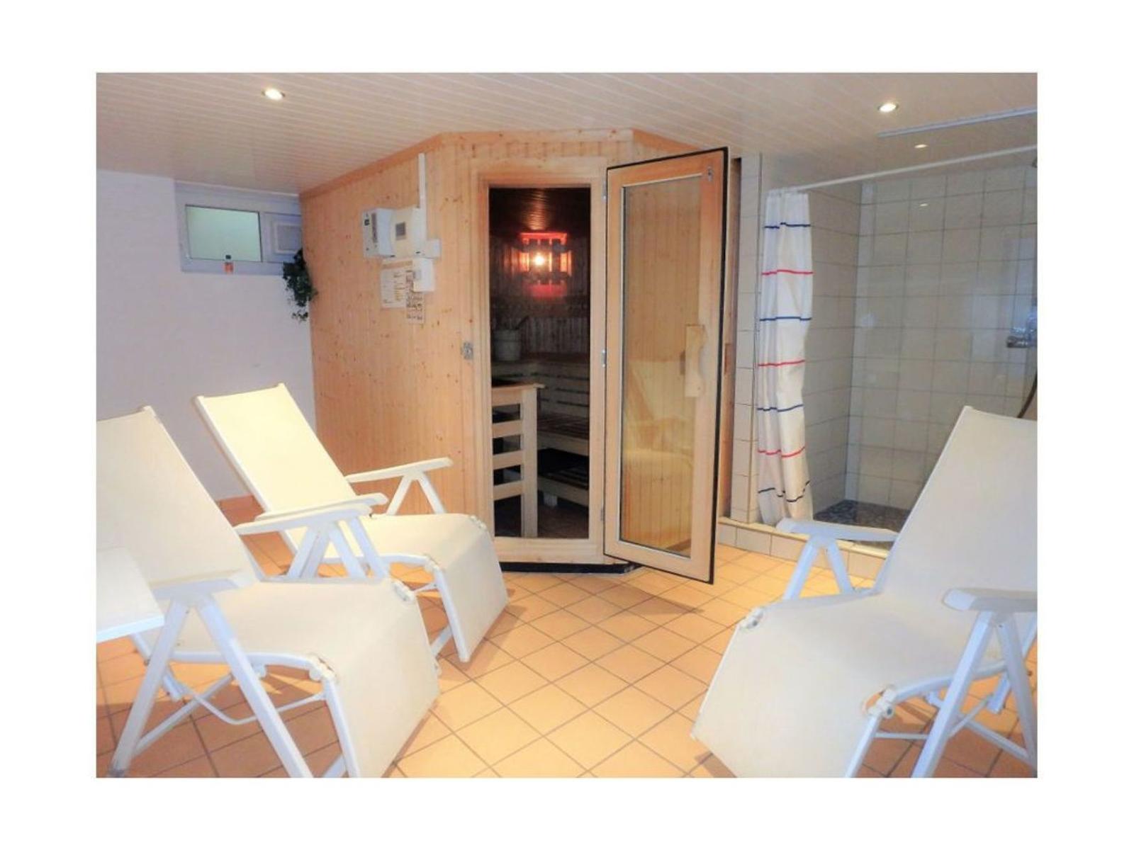Sauna mit Holzinnenraum und drei weißen Liegestühlen. Duschbereich mit weißen Fliesen und Duschvorhang.