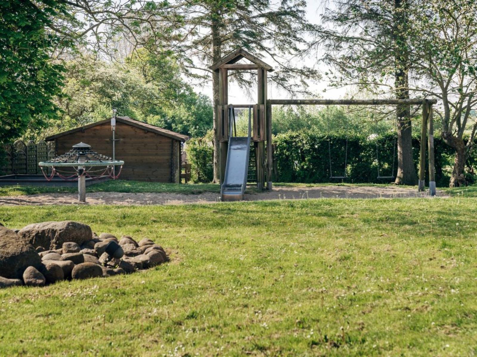 Grüner Garten mit Holzspielplatz, Rutsche und Schaukel. Im Hintergrund steht eine Holzhütte.
