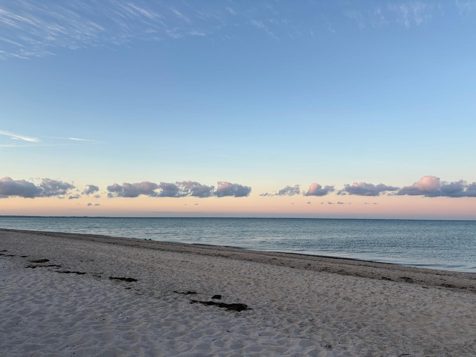 Strand mit feinem Sand und ruhigem Meer unter einem klaren Himmel mit rosa gefärbten Wolken.