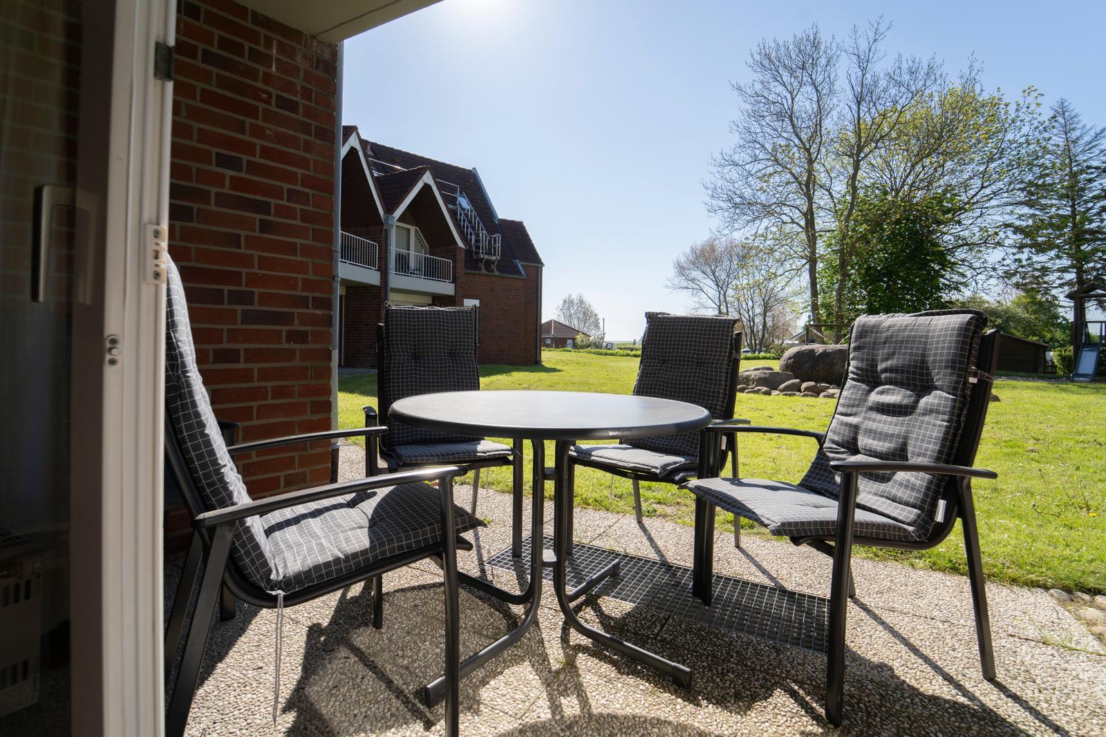 Terrasse mit Tisch und vier Stühlen. Hintergrund: Grasfläche, Bäume und Gebäude.