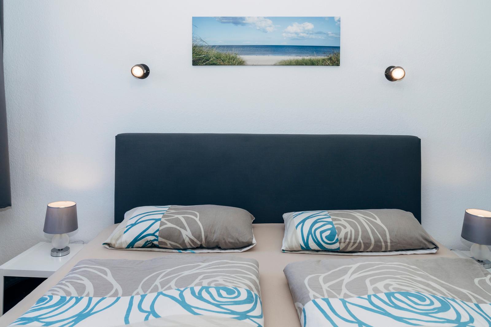Doppelbett mit Kopfkissen und Bettwäsche mit Blumenmuster. Wandbild und zwei Nachttische mit Lampen.