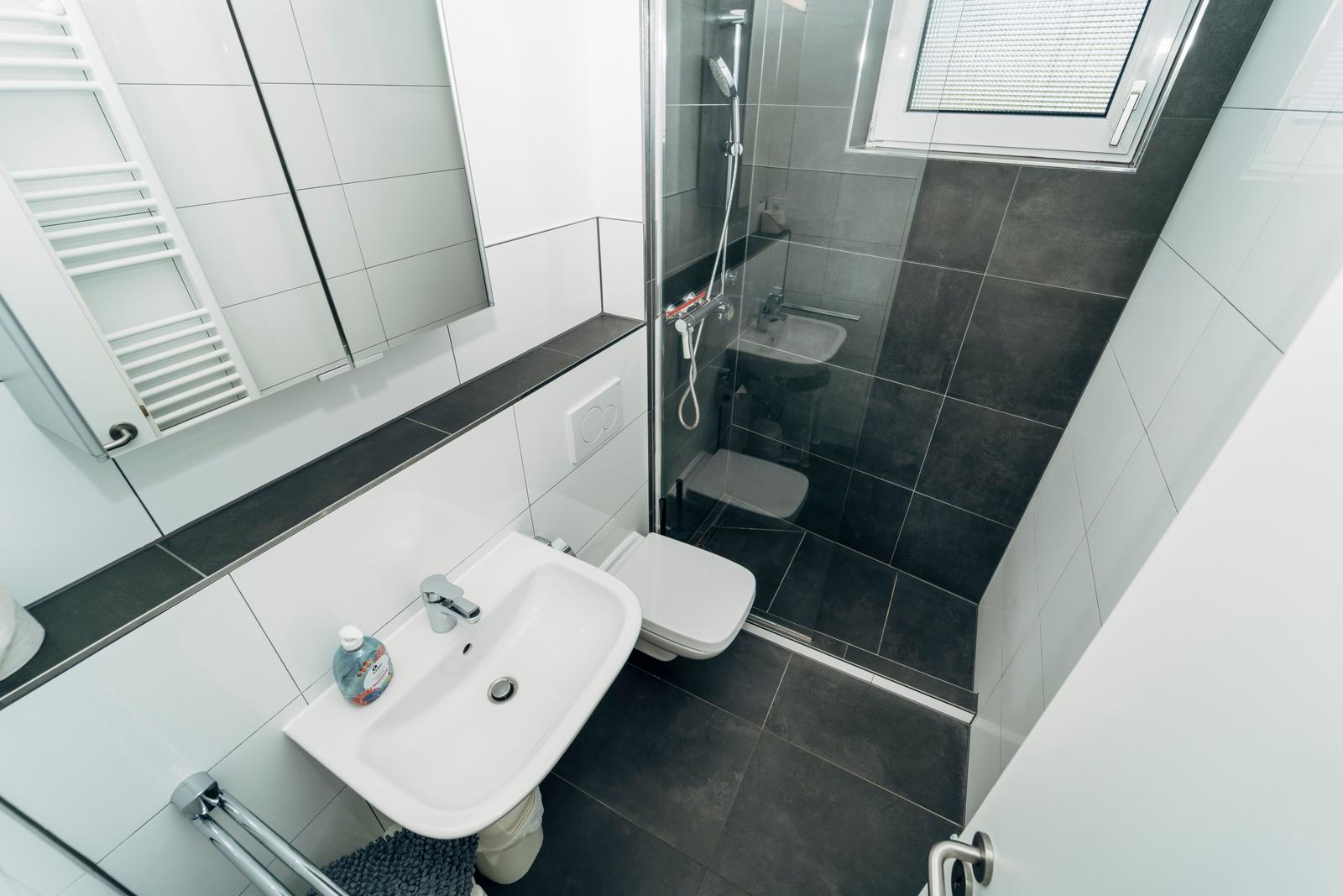 Badezimmer mit Dusche, Waschbecken und Toilette. Fliesen in Grau und Weiß.