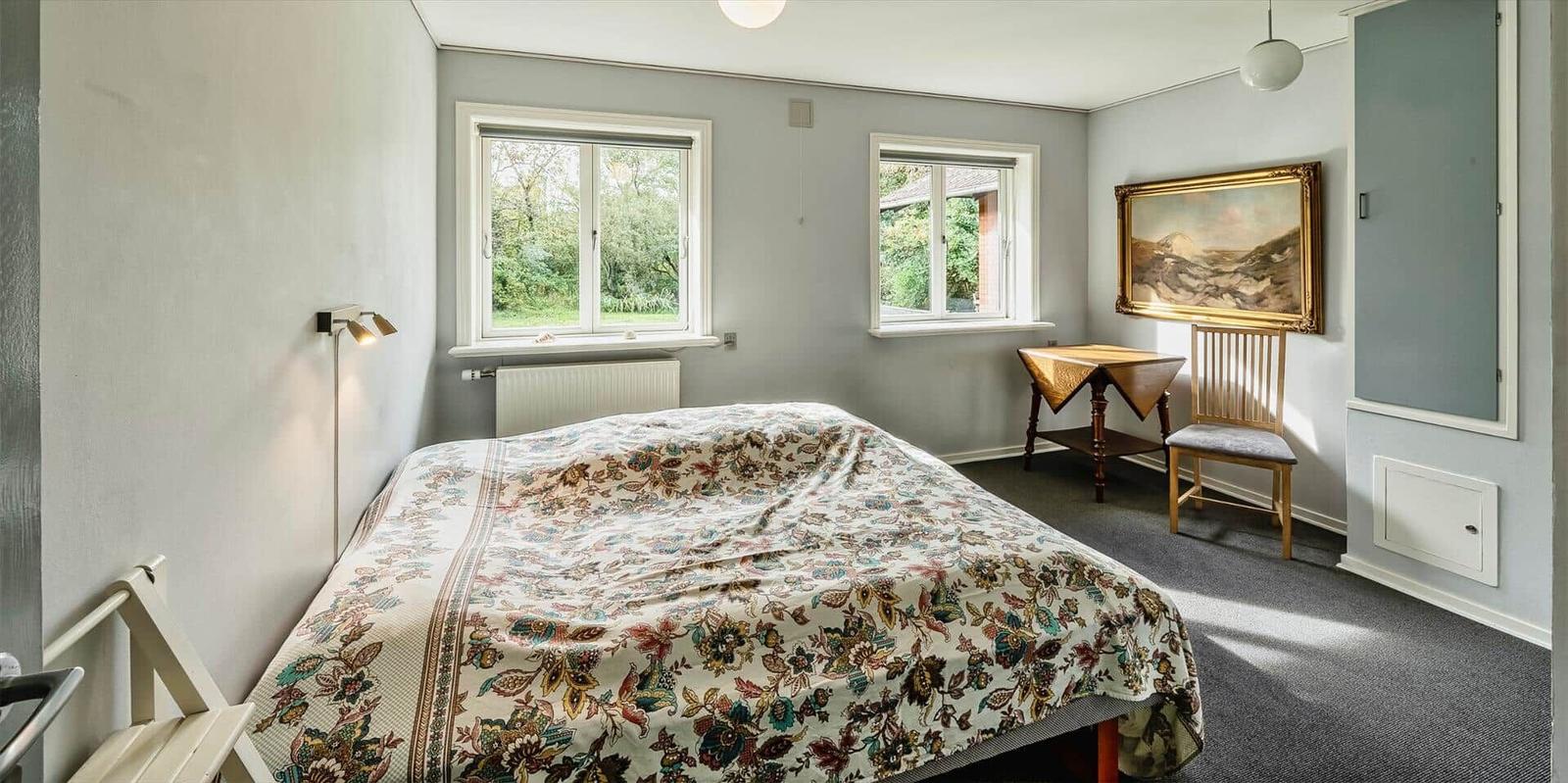 Schlafzimmer mit Bett, zwei Fenstern, Tisch und Stuhl.