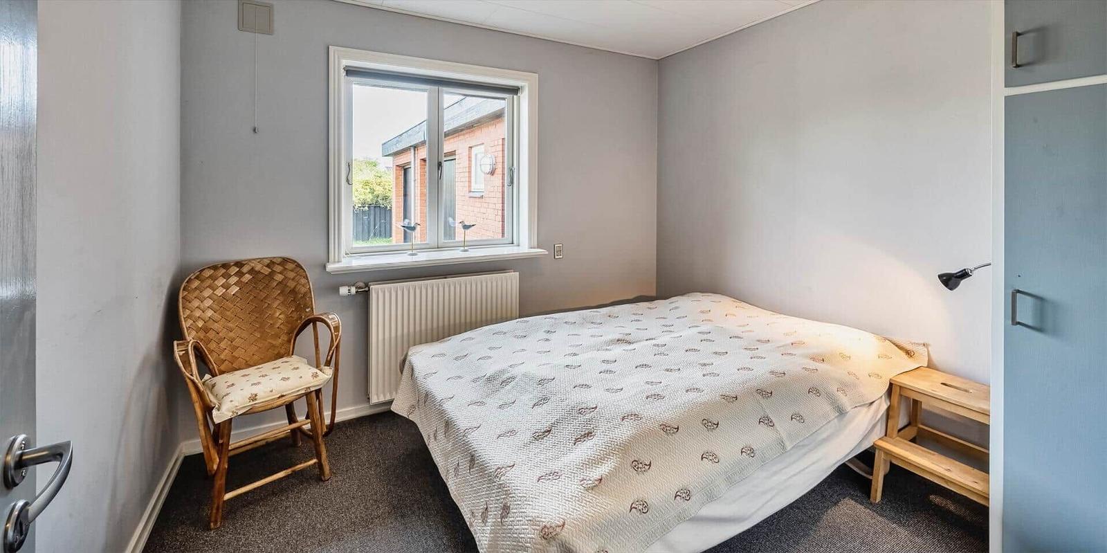 Schlafzimmer mit Bett, Stuhl und Fenster. An der Wand hängt eine Lampe.