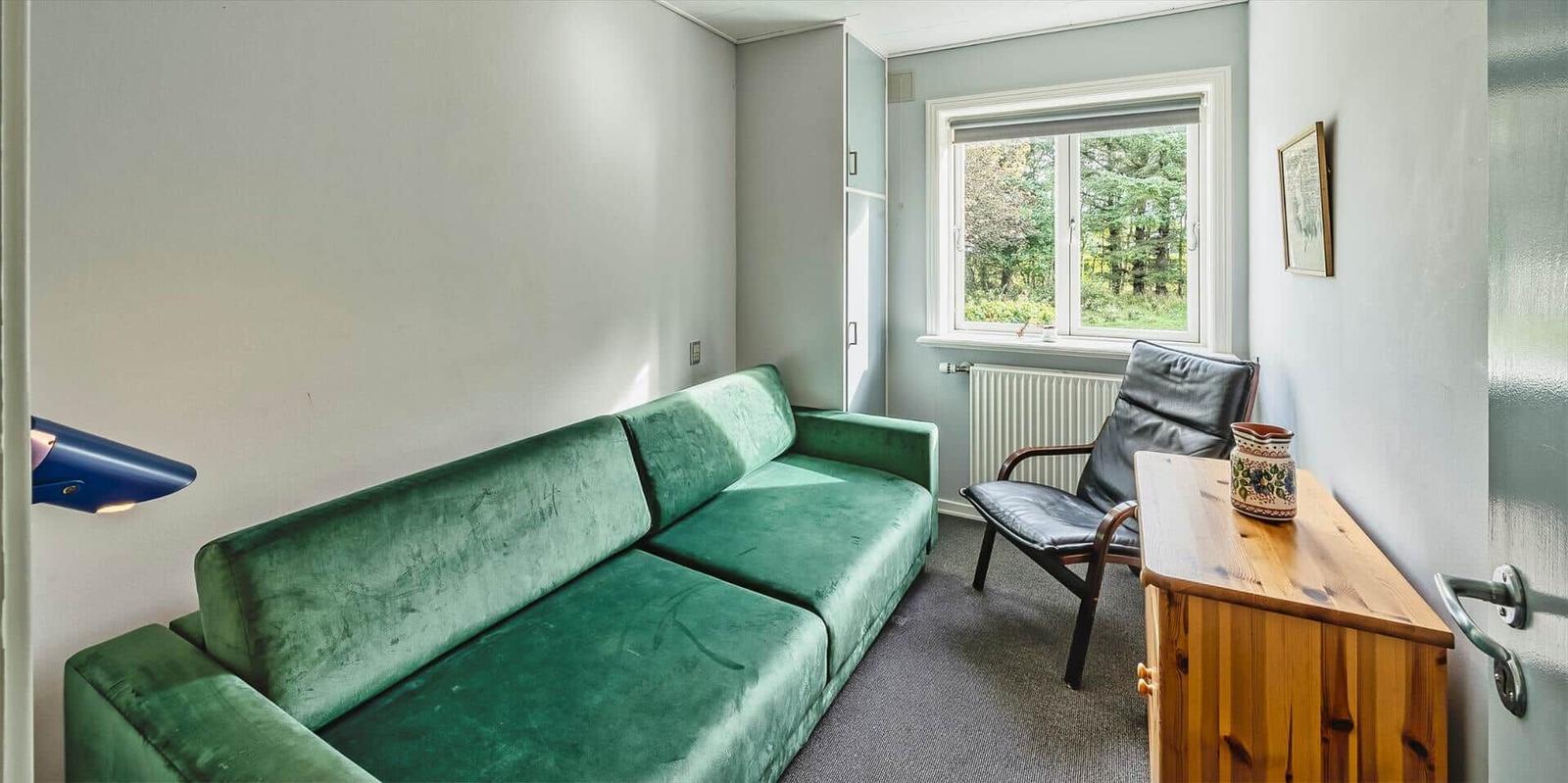 Ein Raum mit grünem Sofa, Stuhl und Holztisch. Fenster mit Blick auf Wald.