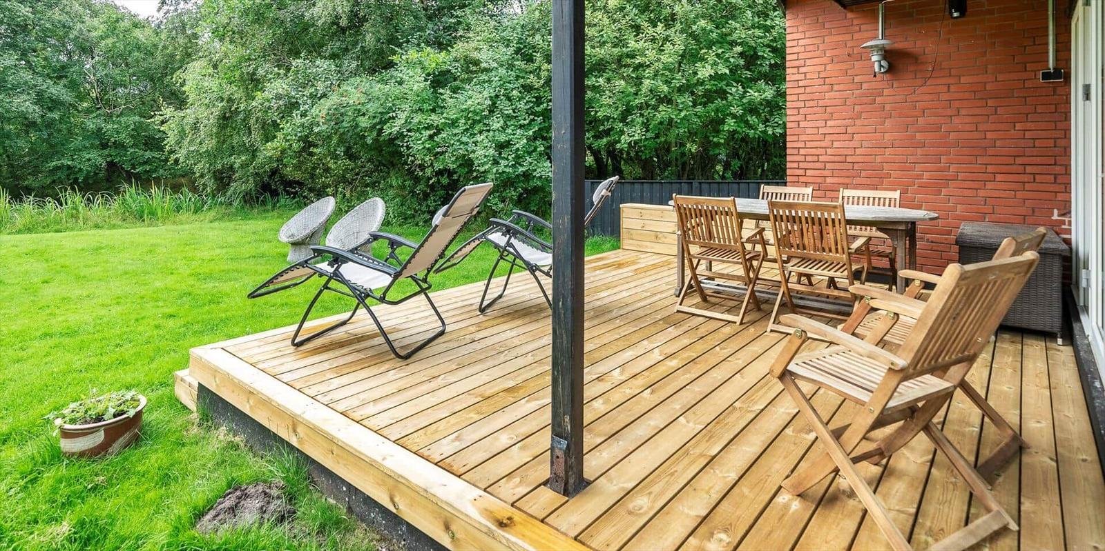 Holzterrasse mit Liegestühlen und Esstisch neben rotem Backsteinhaus.