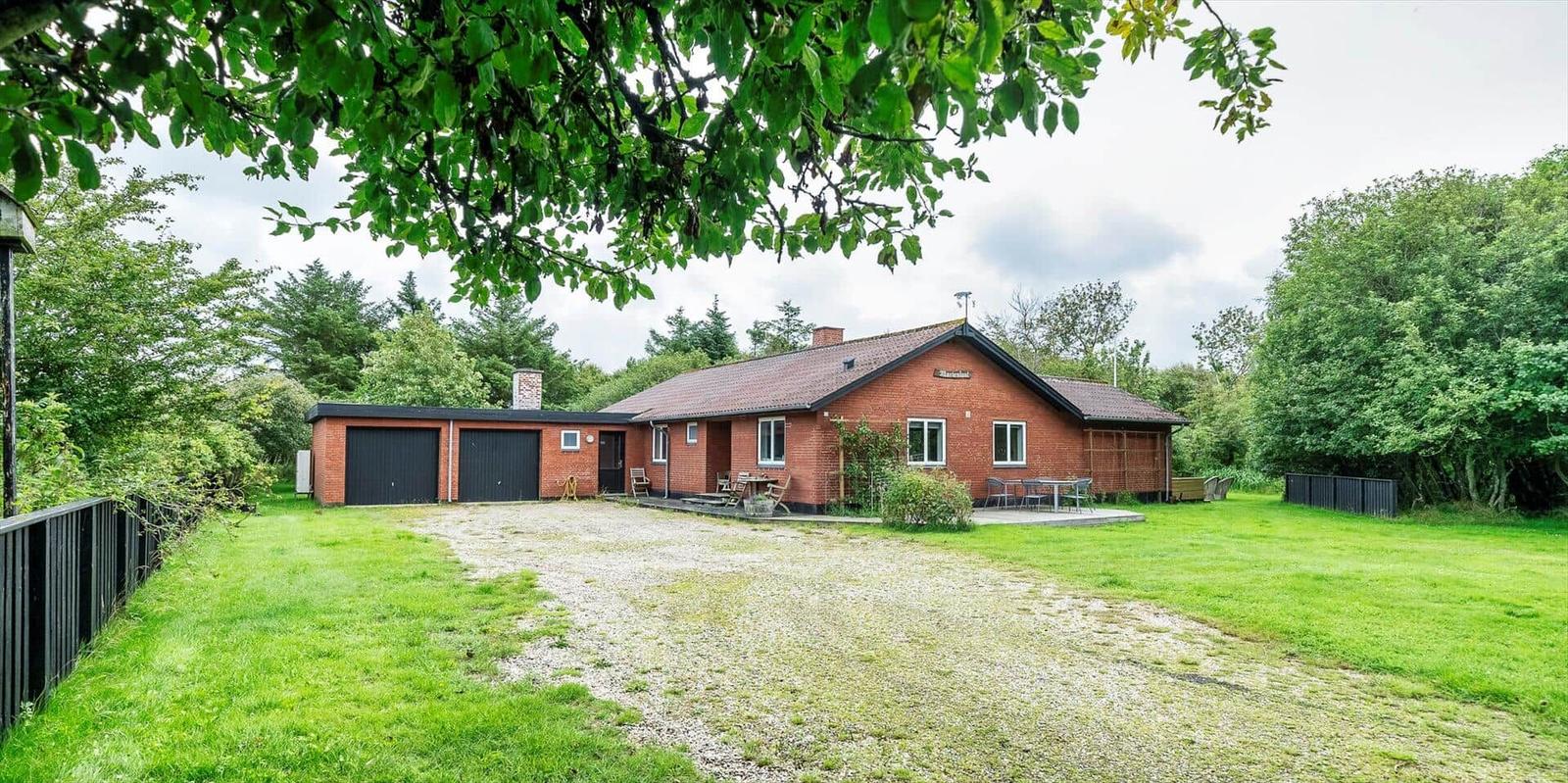 Rotes Haus mit Garage, Terrasse und Garten. Umgeben von Bäumen und Gras.