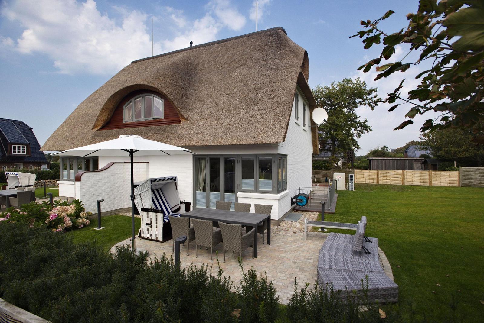 Haus mit Strohdach, Terrasse mit Tisch, Garten und Sonnenschirm.