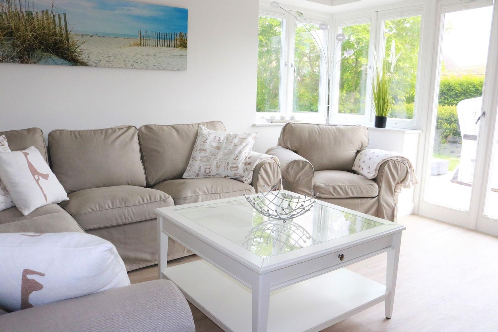 Wohnzimmer mit Sofa, Sessel und Glas-Tisch. Große Fenster und Wandbild mit Strandmotiv.