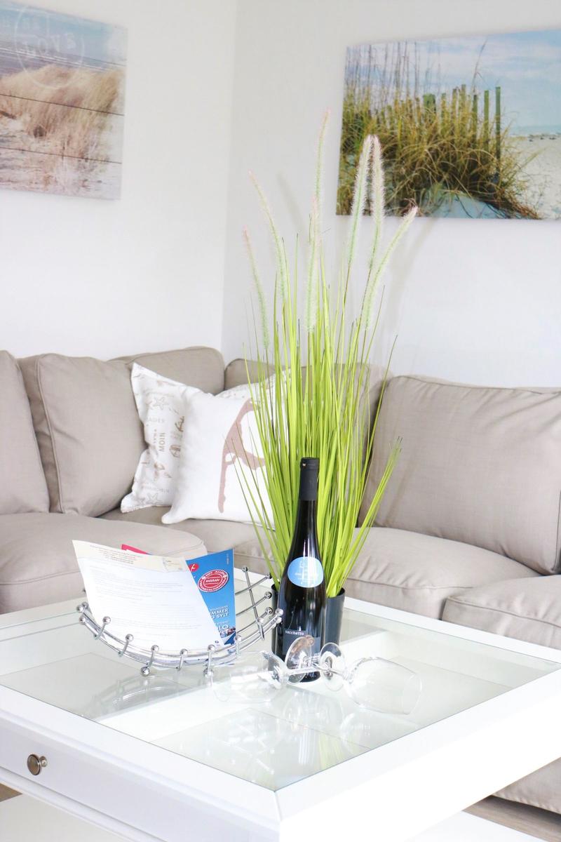 Glas-Tisch mit Flasche, Gläsern und Pflanze vor einem Sofa.