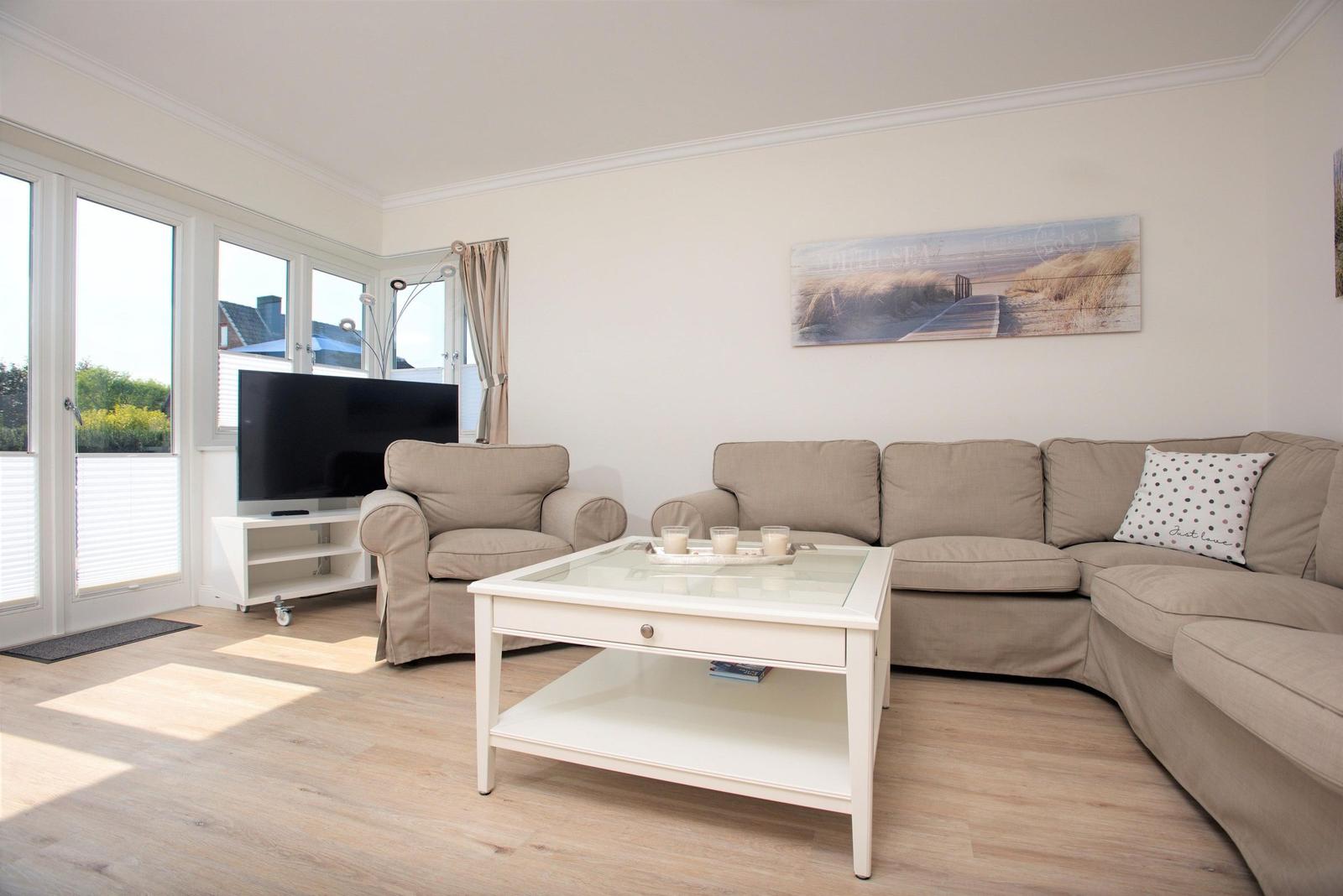 Wohnzimmer mit Sofa, Sessel, Glas-Tisch und TV. Fenster mit Blick auf Außenbereich.