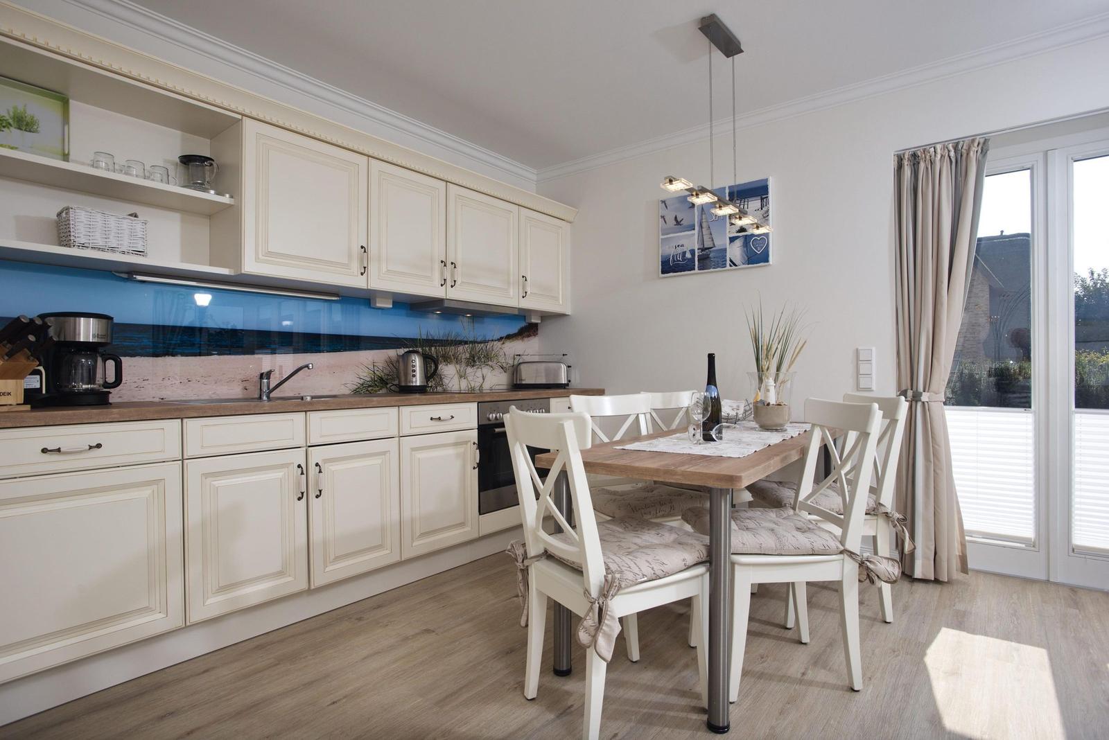Küche mit Essbereich, weiße Schränke, Holztisch und Stühle, blaue Rückwand und Fenster mit Vorhängen.