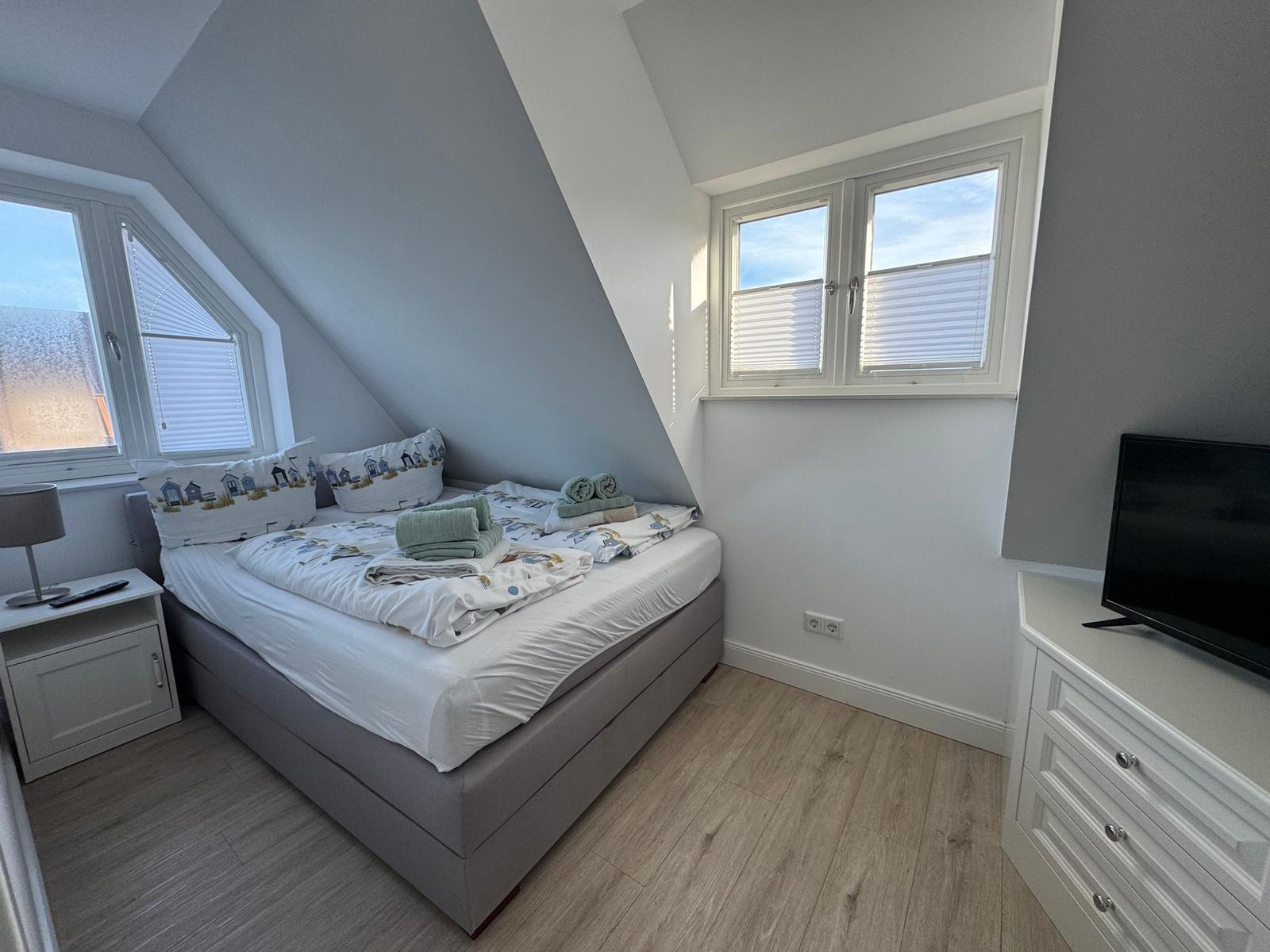 Schlafzimmer mit Dachschrägen, Bett, TV und Fenstern.