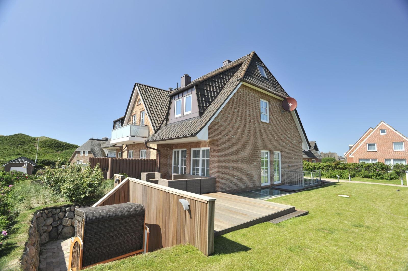 Modernes Haus mit Holzterrasse und Garten. Satellitenschüssel und Fenster sichtbar.