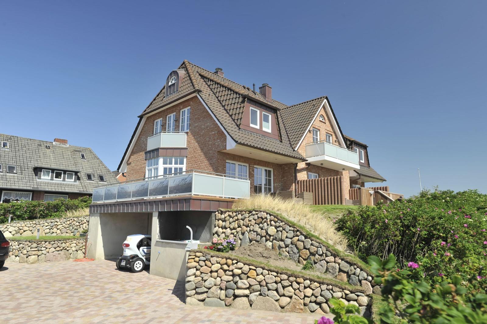 Zweistöckiges Haus mit Balkon und Garage. Steinmauer und Grünanlagen.
