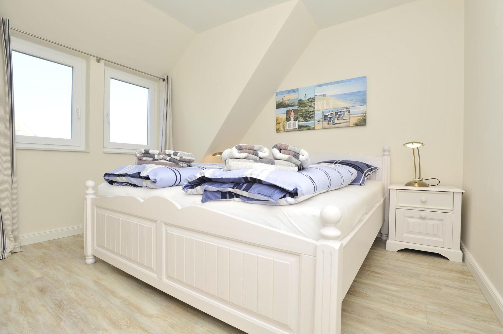 Schlafzimmer mit weißem Bett, zwei Fenstern und Nachttisch mit Lampe.