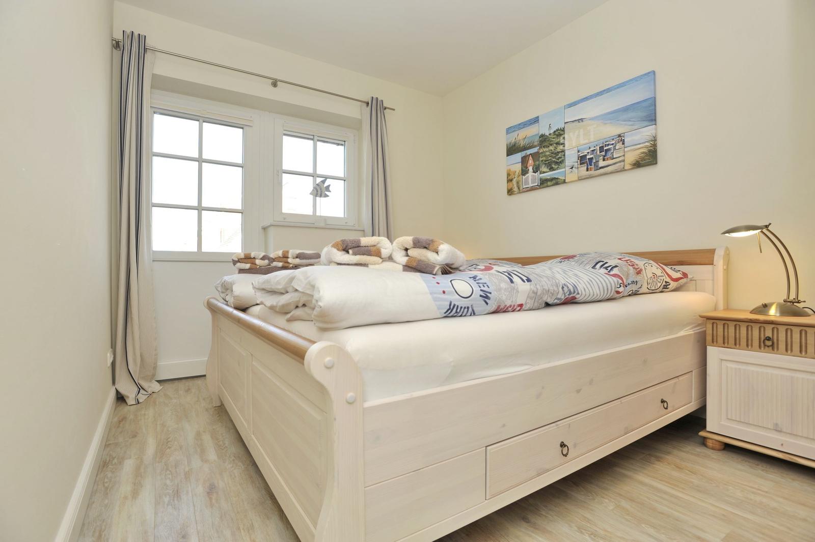 Schlafzimmer mit Bett, Nachttisch und Fenster. Wandbild zeigt Strand.