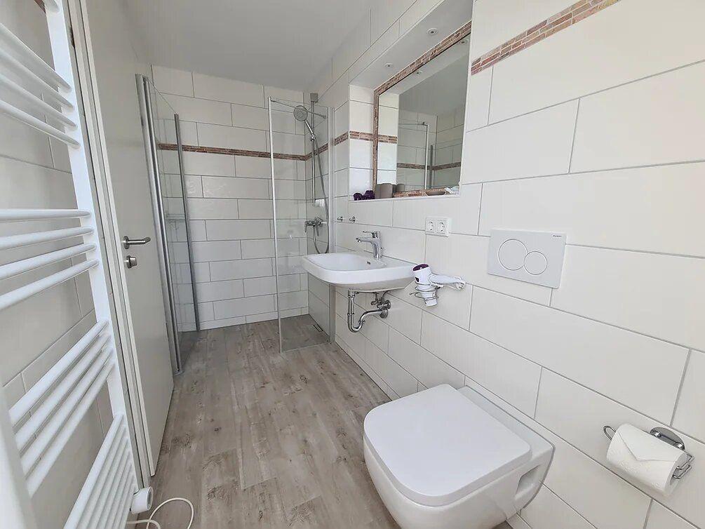 Bathroom with shower, sink, and toilet. White tiles and wooden floor.