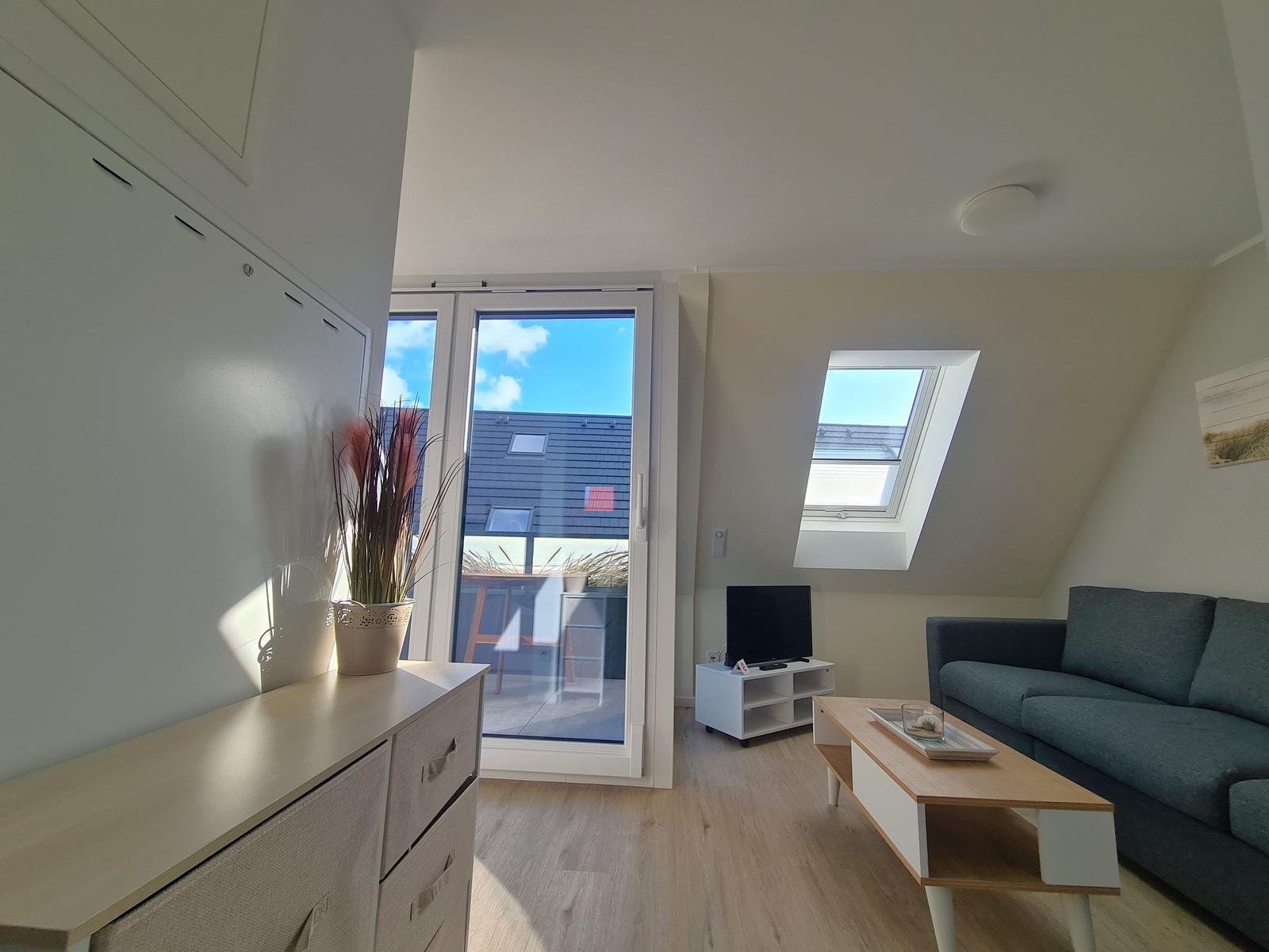 Wohnzimmer mit Sofa, TV und Balkonzugang. Holzboden und Dachfenster.