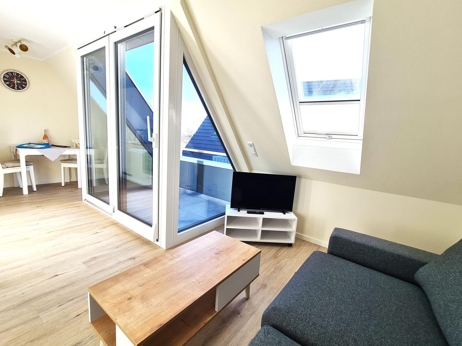 Wohnzimmer mit Sofa, TV und Balkontür. Holzdielen und Fenster.