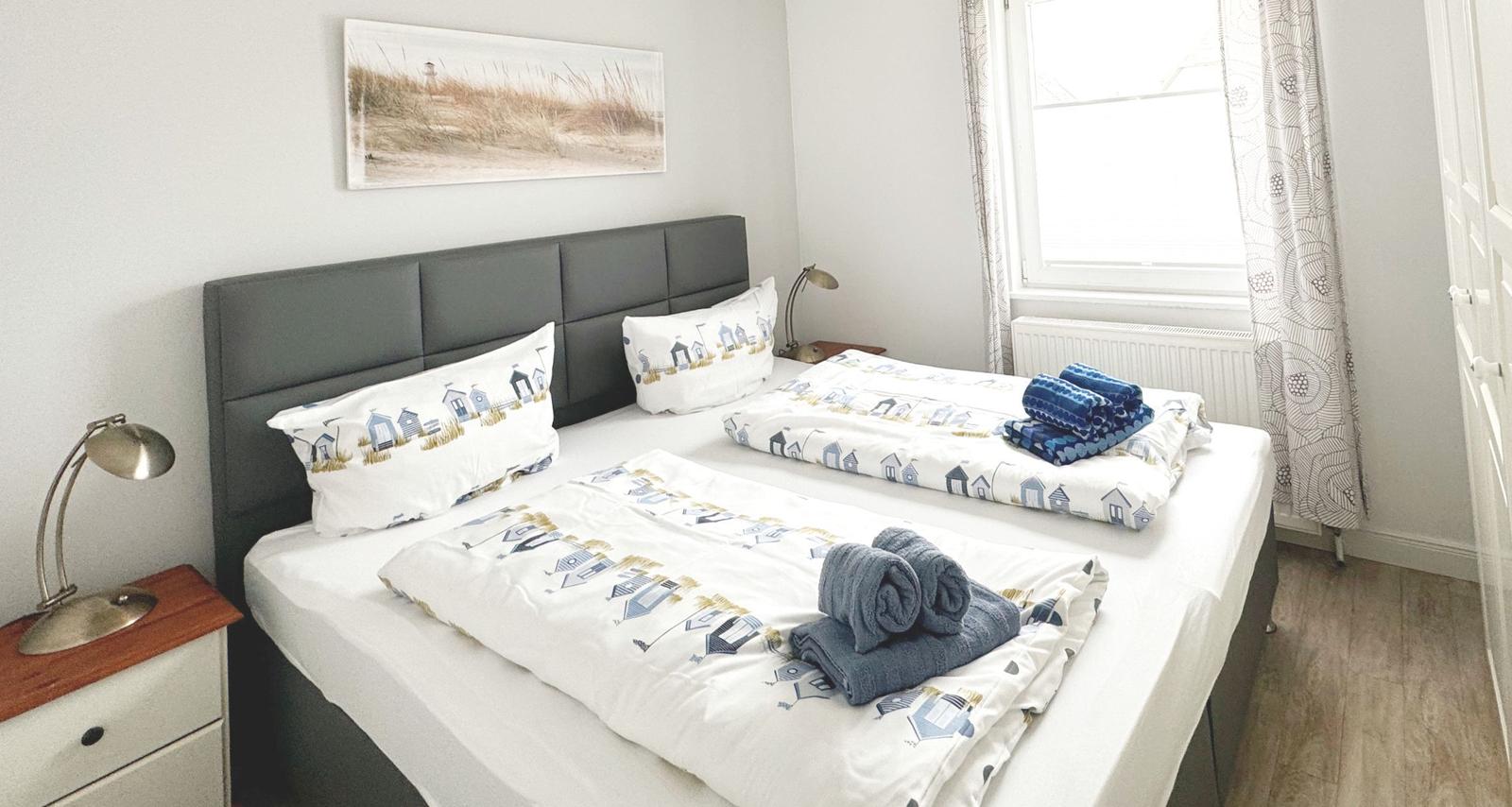 Schlafzimmer mit Doppelbett, Bettwäsche mit Strandmotiv und Fenster mit Vorhängen.