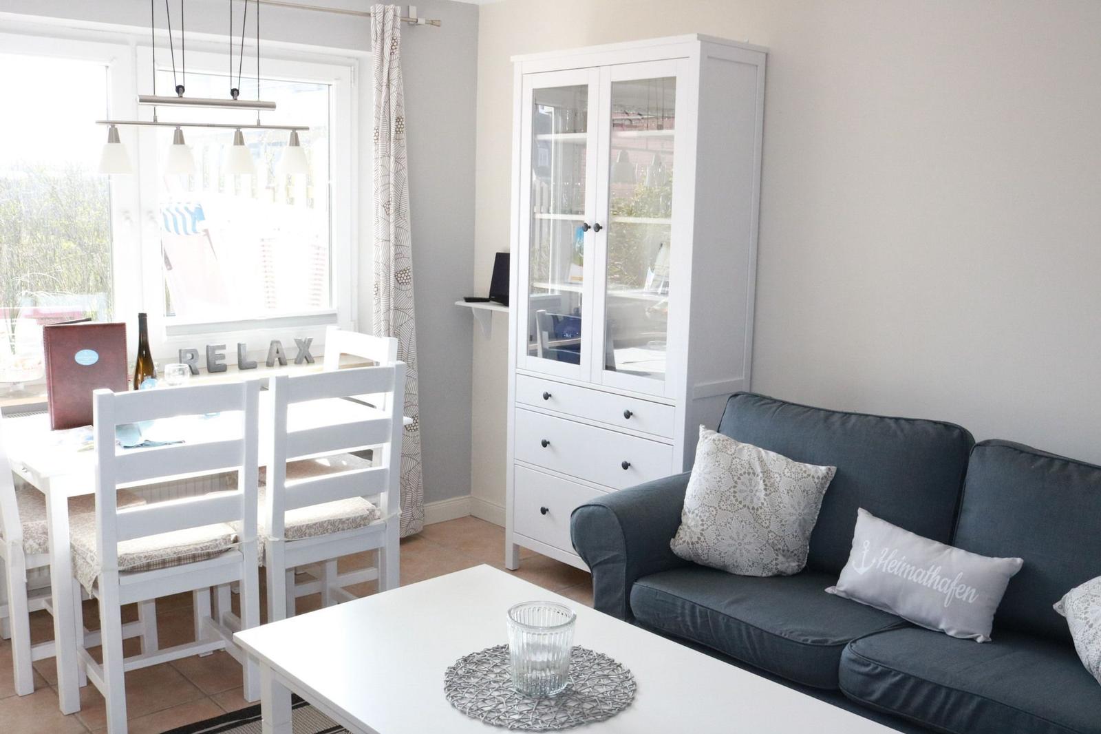Living and dining area with sofa, table, and white chairs. Background: window with curtains.