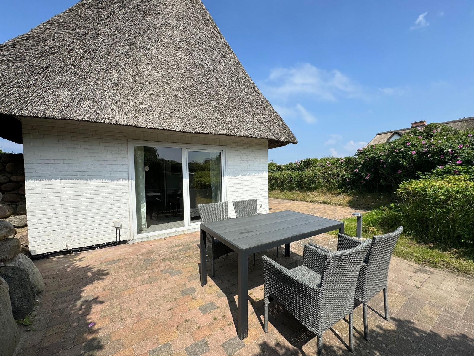 Terrace with table and chairs in front of a house with thatched roof.