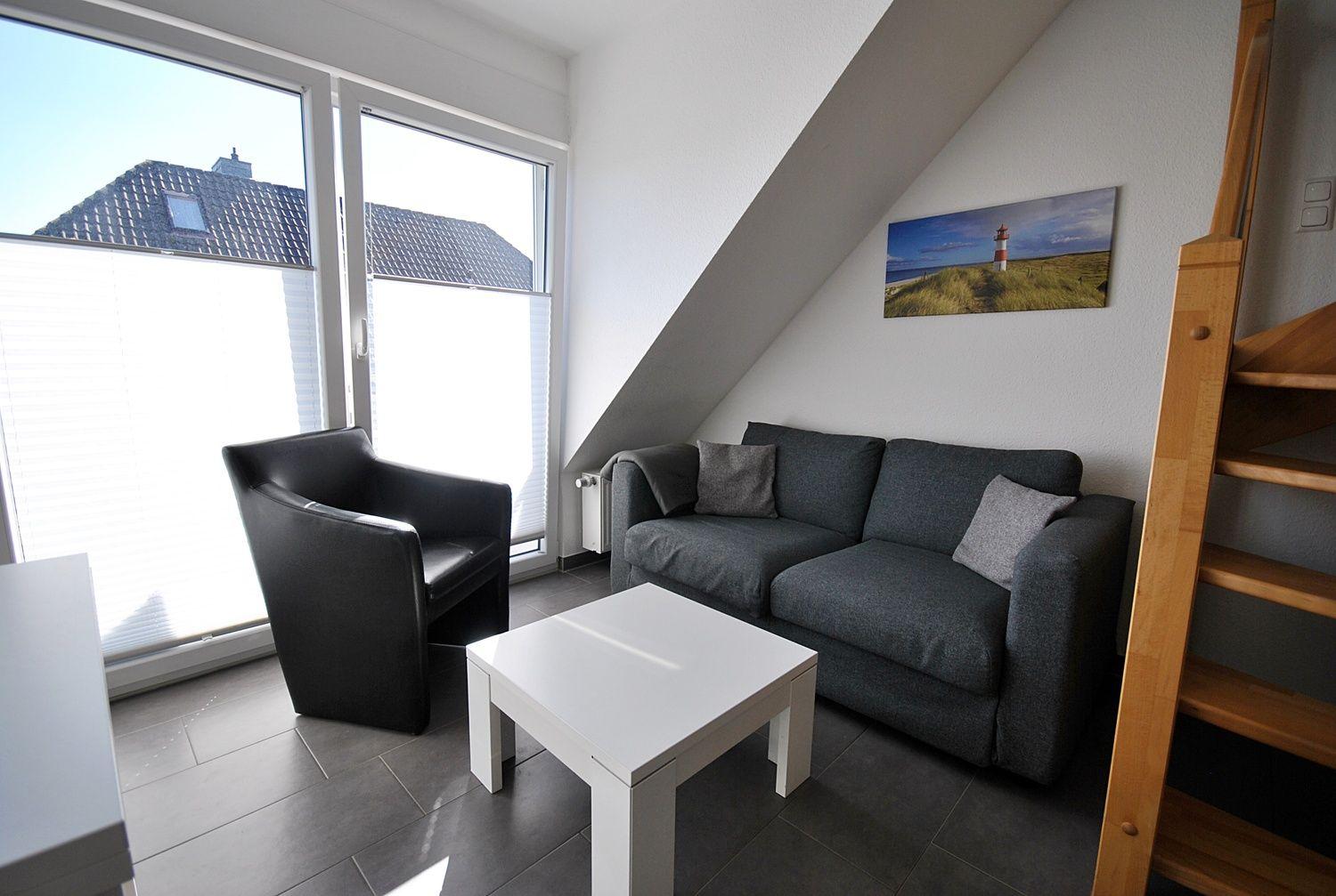 Wohnzimmer mit Sofa, Sessel und Tisch. Fenster mit Blick auf Dach. Holztreppe und Wandbild.