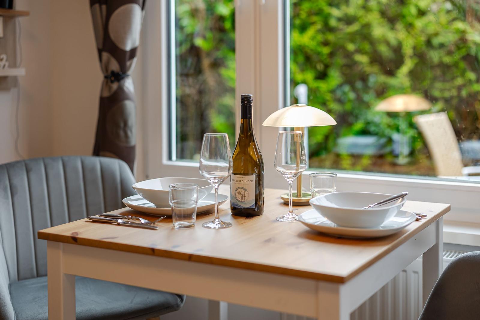 Esstisch mit Geschirr, Weinflasche und Lampe vor Fenster mit Blick auf Garten.