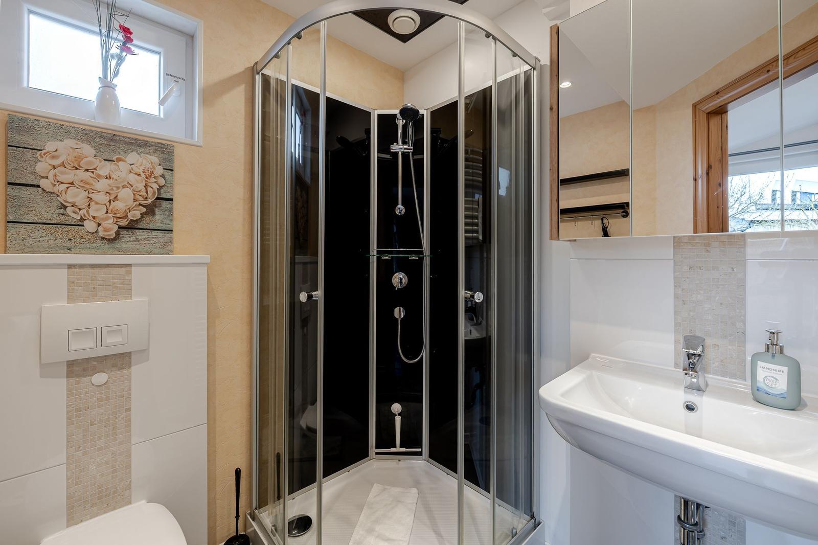Badezimmer mit Dusche, Waschbecken und Toilette. Fenster, Spiegel und Wanddekoration sichtbar.