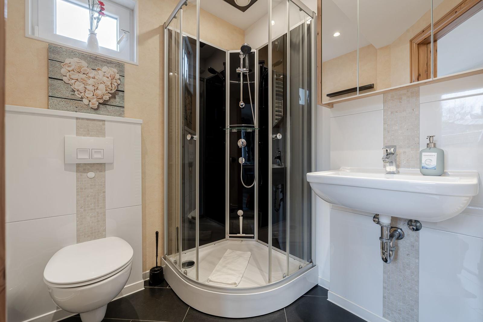 Badezimmer mit Dusche, Waschbecken und Toilette. Wandbild und Fenster sichtbar.