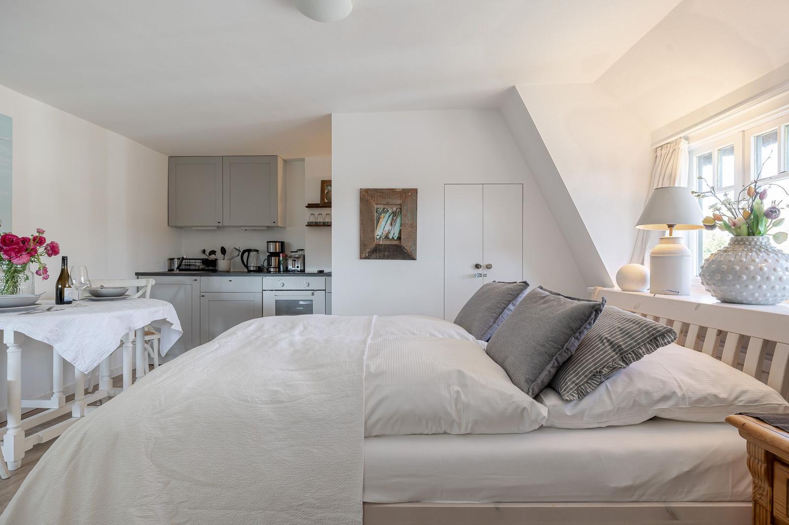 Bedroom with view of kitchen and window. Bed, table, and cabinets are visible.
