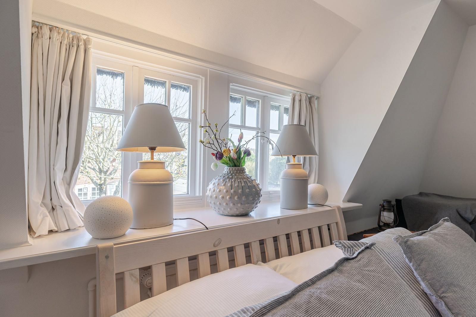 Bedroom with windows, lamps, and vase on windowsill.