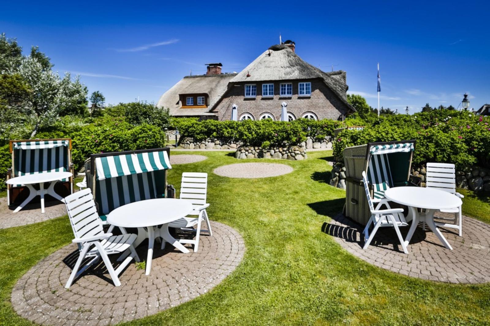 Garten mit Liegestühlen und Tischen vor einem strohgedeckten Haus.