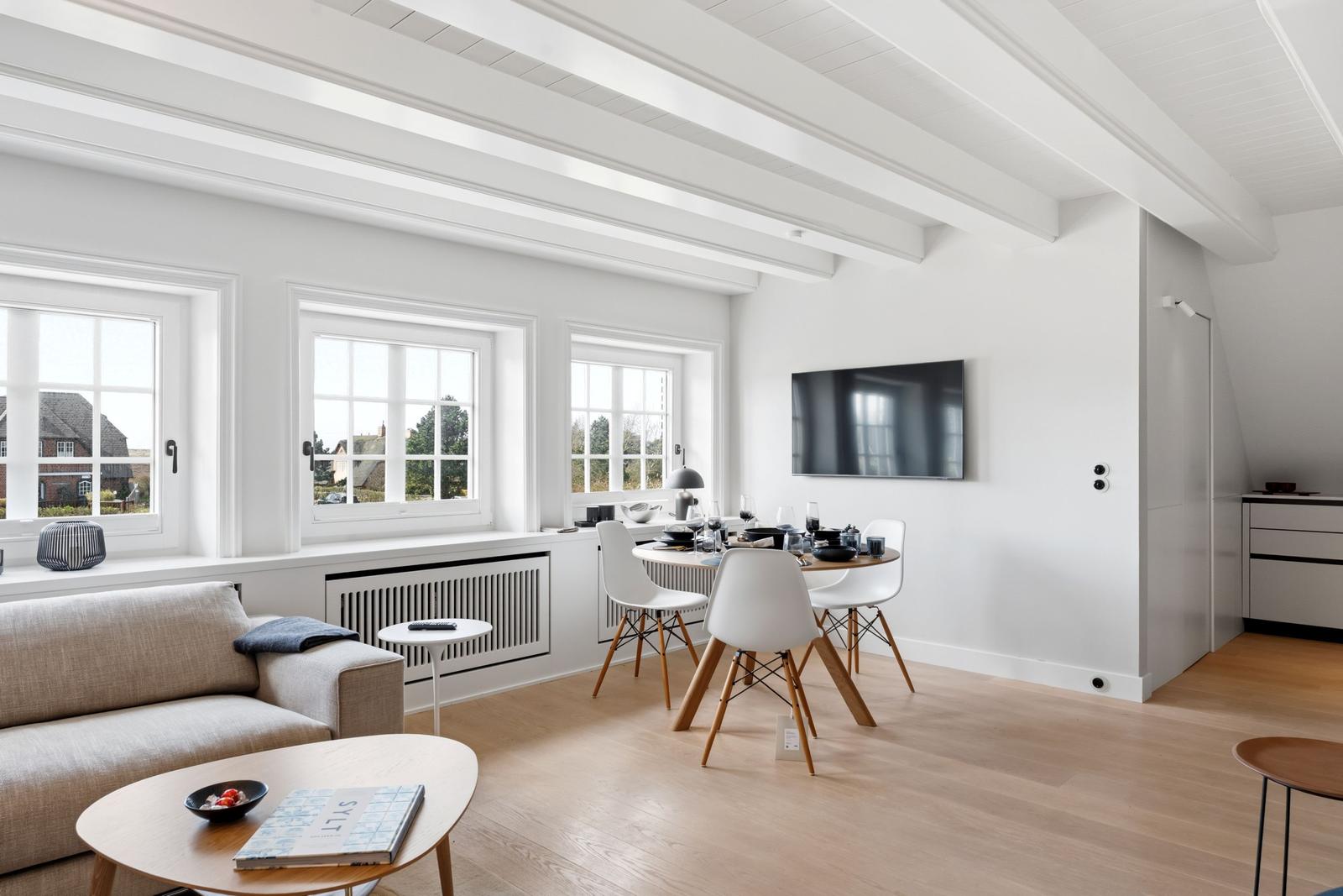 Wohn- und Essbereich mit weißen Wänden, Holzboden und Fenstern. Esstisch mit Stühlen und Fernseher.