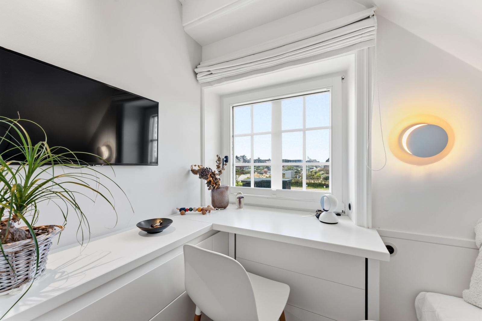 Schreibtisch mit Fensterblick, Wand-TV und Pflanze.