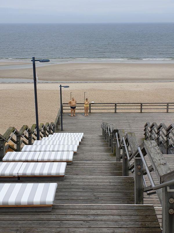 Holzterrasse mit Liegestühlen führt zum Strand. Zwei Personen stehen am Geländer.