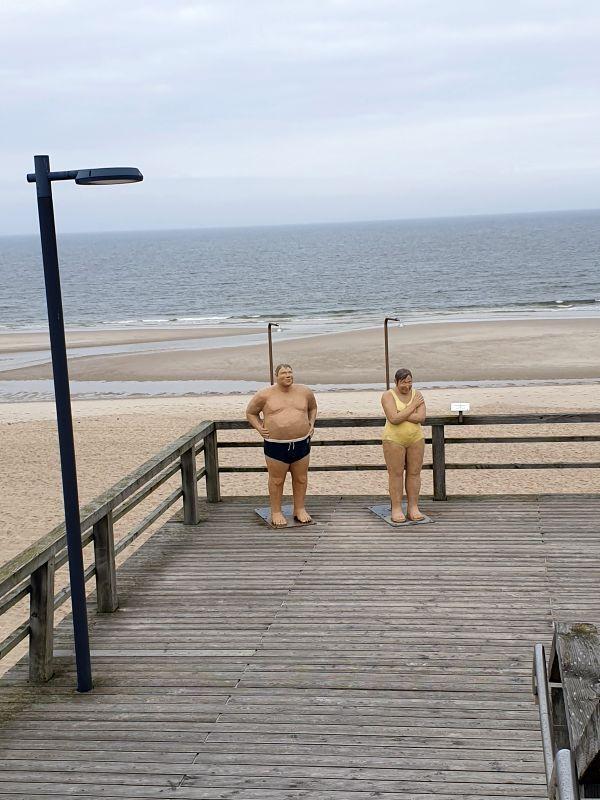 Zwei Figuren stehen auf einer Holzterrasse mit Blick auf den Strand und das Meer.