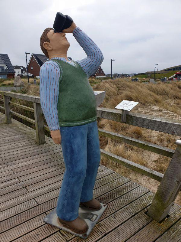 Statue eines Mannes mit Brille auf Holzbrücke vor Dünen