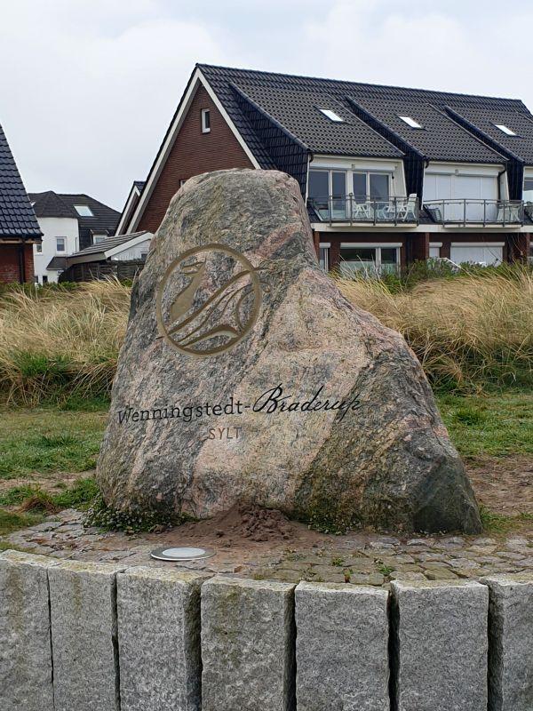 Großer Stein mit Aufschrift Wenningstedt-Braderup, Sylt, vor Wohnhäusern mit Balkonen.