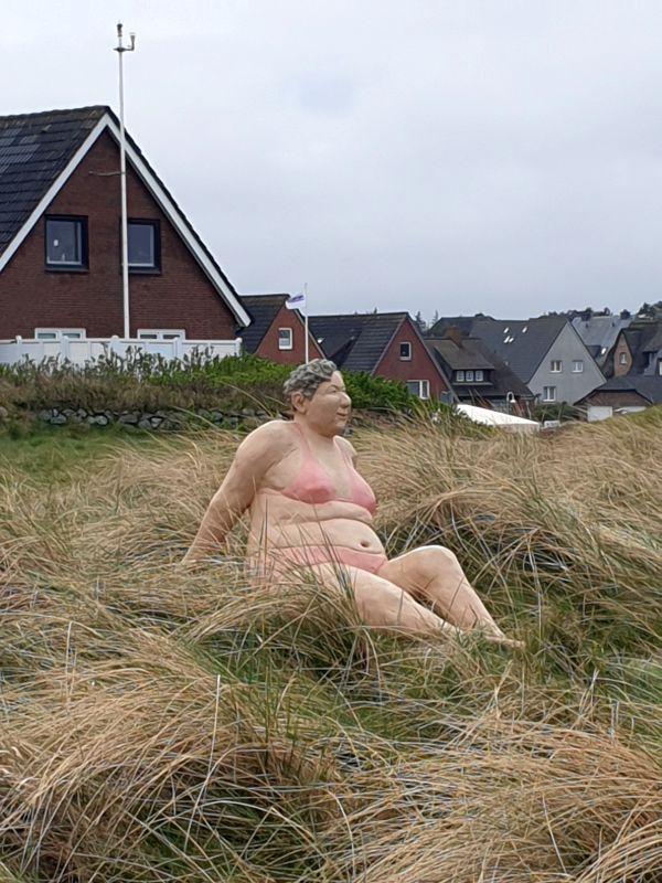 Eine Skulptur einer Frau im Bikini sitzt im hohen Gras. Hintergrund: Wohnhäuser und bewölkte Himmel.