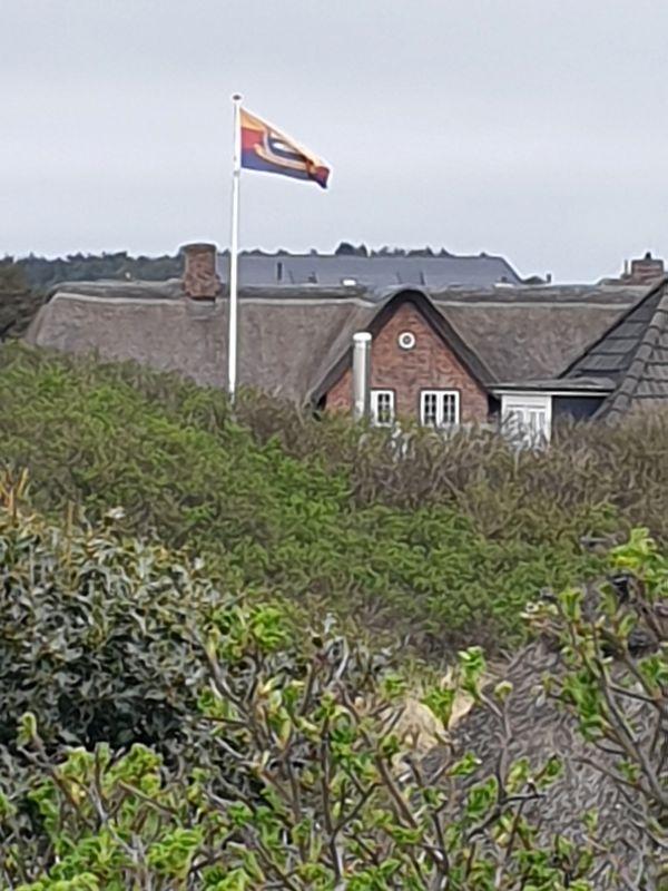 Haus mit Schindeldach und Flagge auf Mast. Umgeben von Grünflächen.