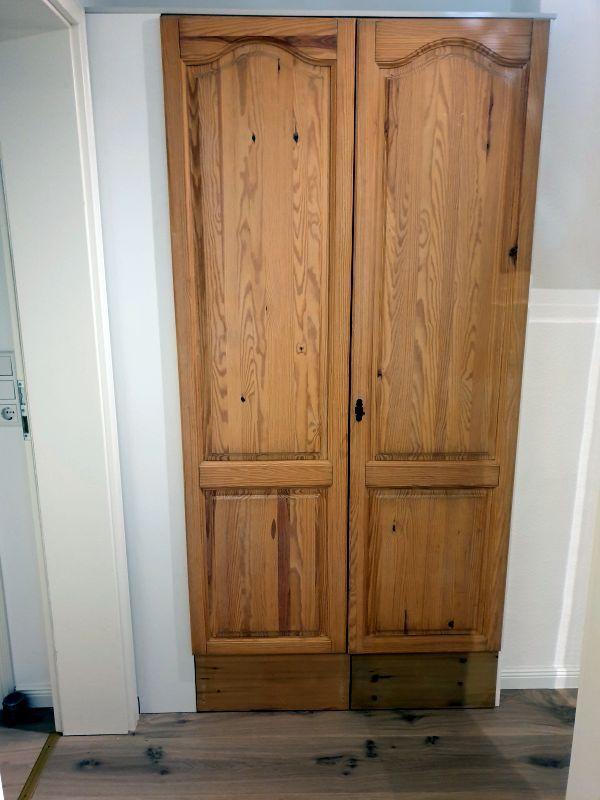 Holzschrank mit zwei Türen und schwarzen Griffen an der Wand.