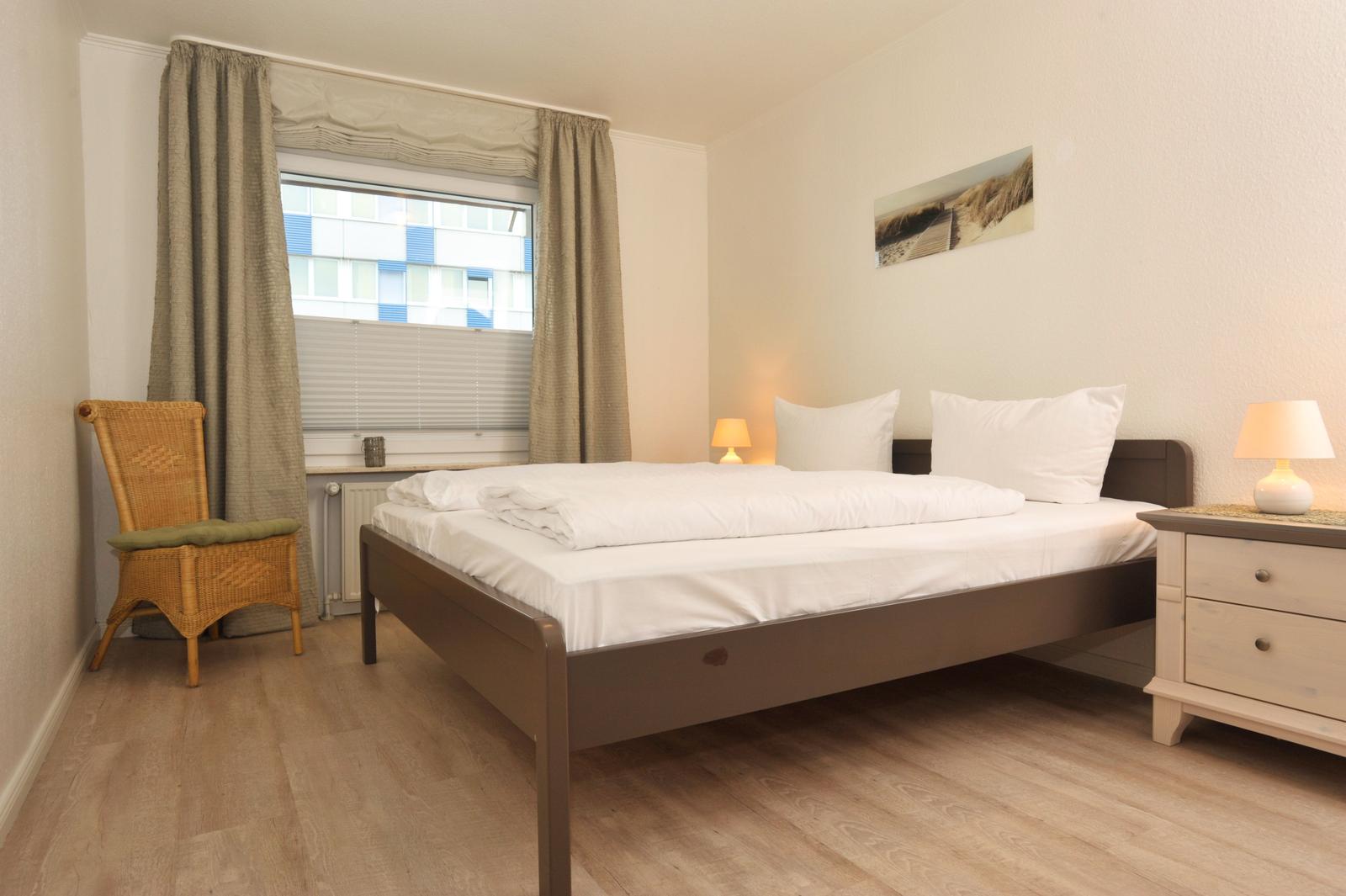 Schlafzimmer mit Bett, Nachttisch und Stuhl. Fenster mit Jalousie und Vorhang.
