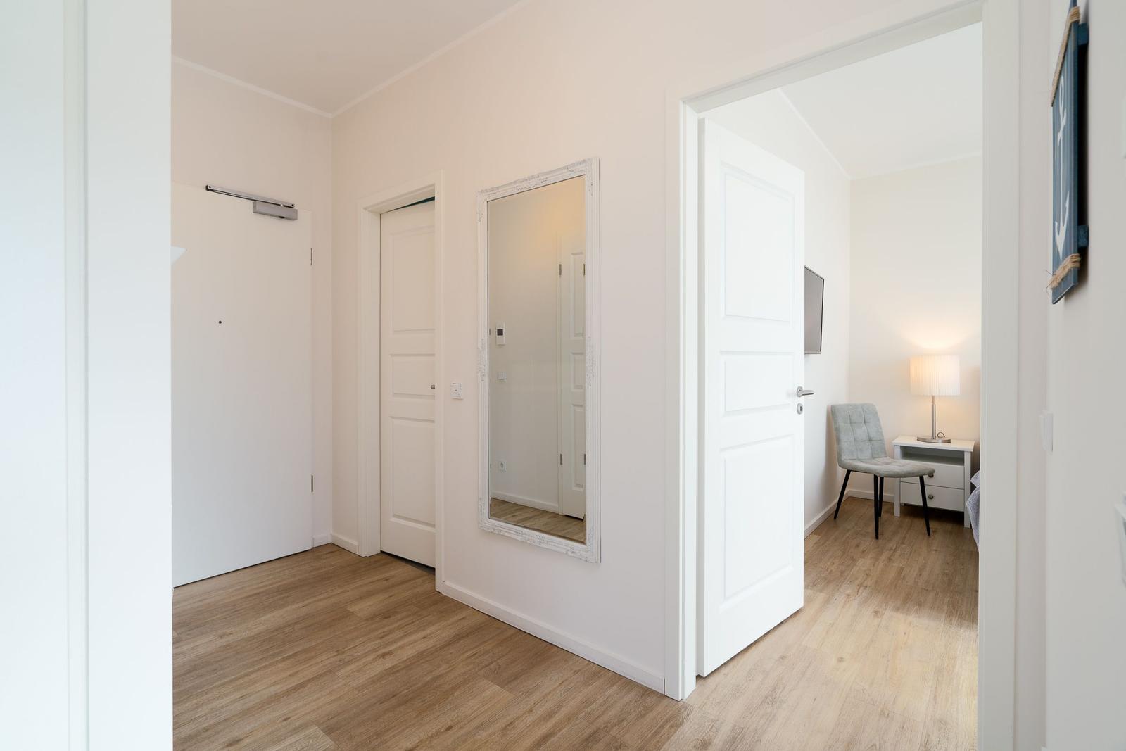 Flur mit Spiegel und Türen. Durch eine Tür ist ein Schlafzimmer mit Stuhl und Nachttisch sichtbar.