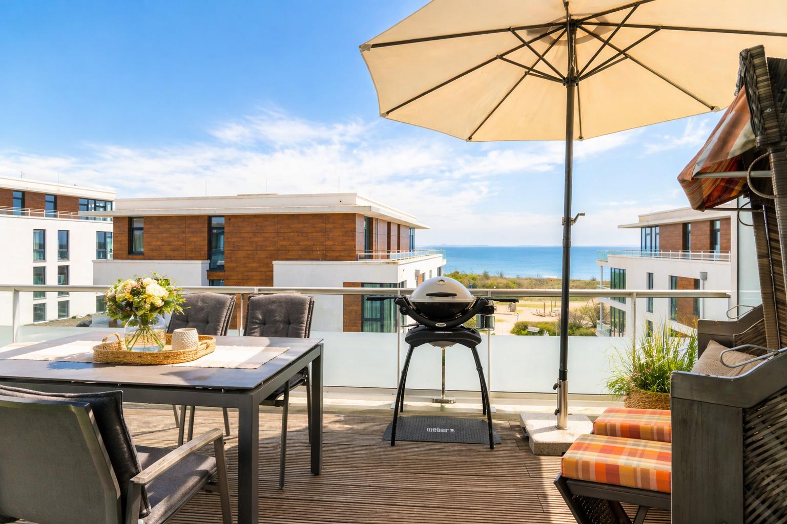 Terrasse mit Tisch, Stühlen, Grill und Sonnenschirm. Blick auf Meer und moderne Gebäude.