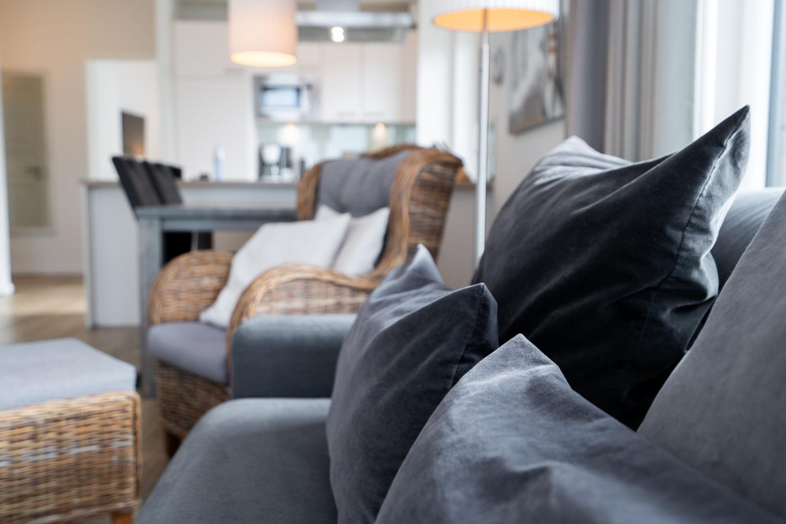 Ein Wohnbereich mit grauem Sofa und Kissen, einem Stuhl aus Rattan und einer Küchenzeile im Hintergrund.