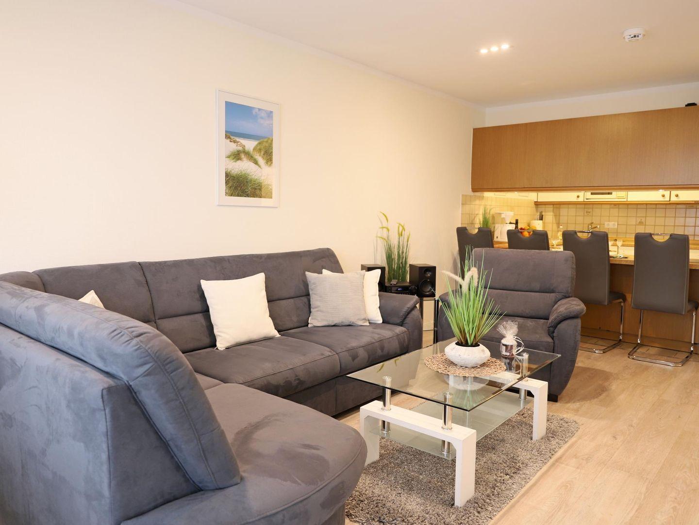 Wohnbereich mit grauem Sofa, Glas-Tisch und Küchenbereich im Hintergrund.