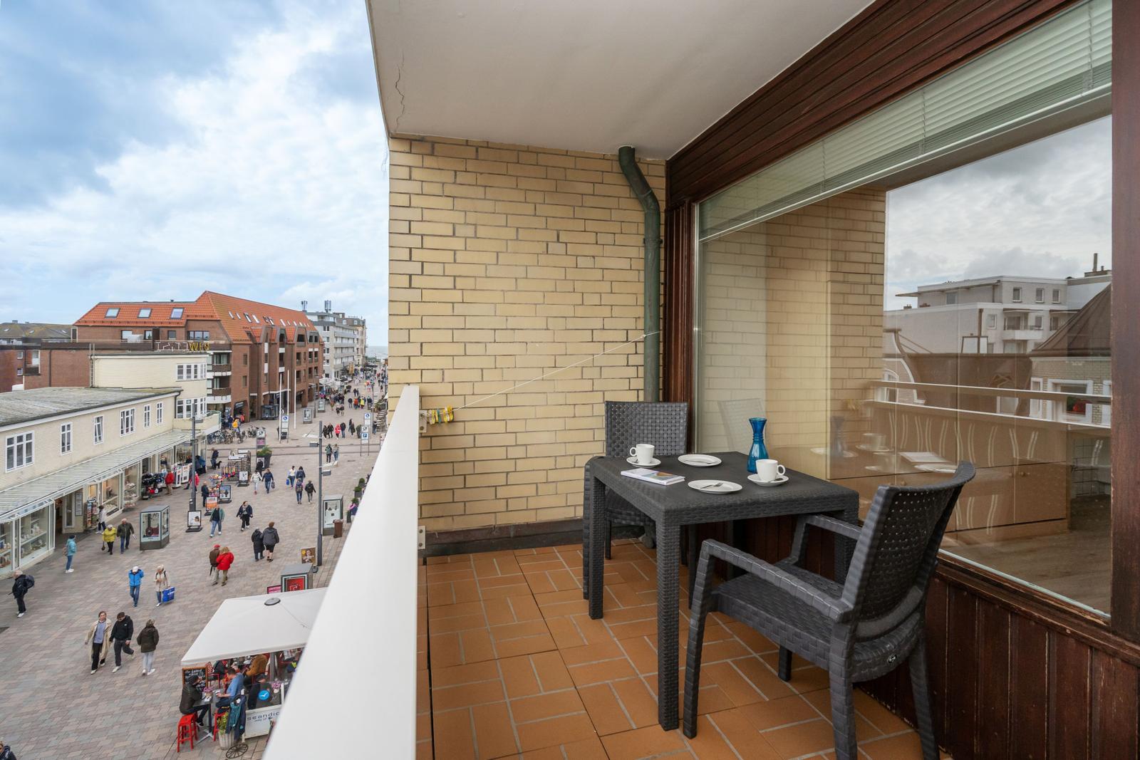 Balkon mit Tisch und Stühlen, Blick auf belebte Straße mit Geschäften und Passanten.