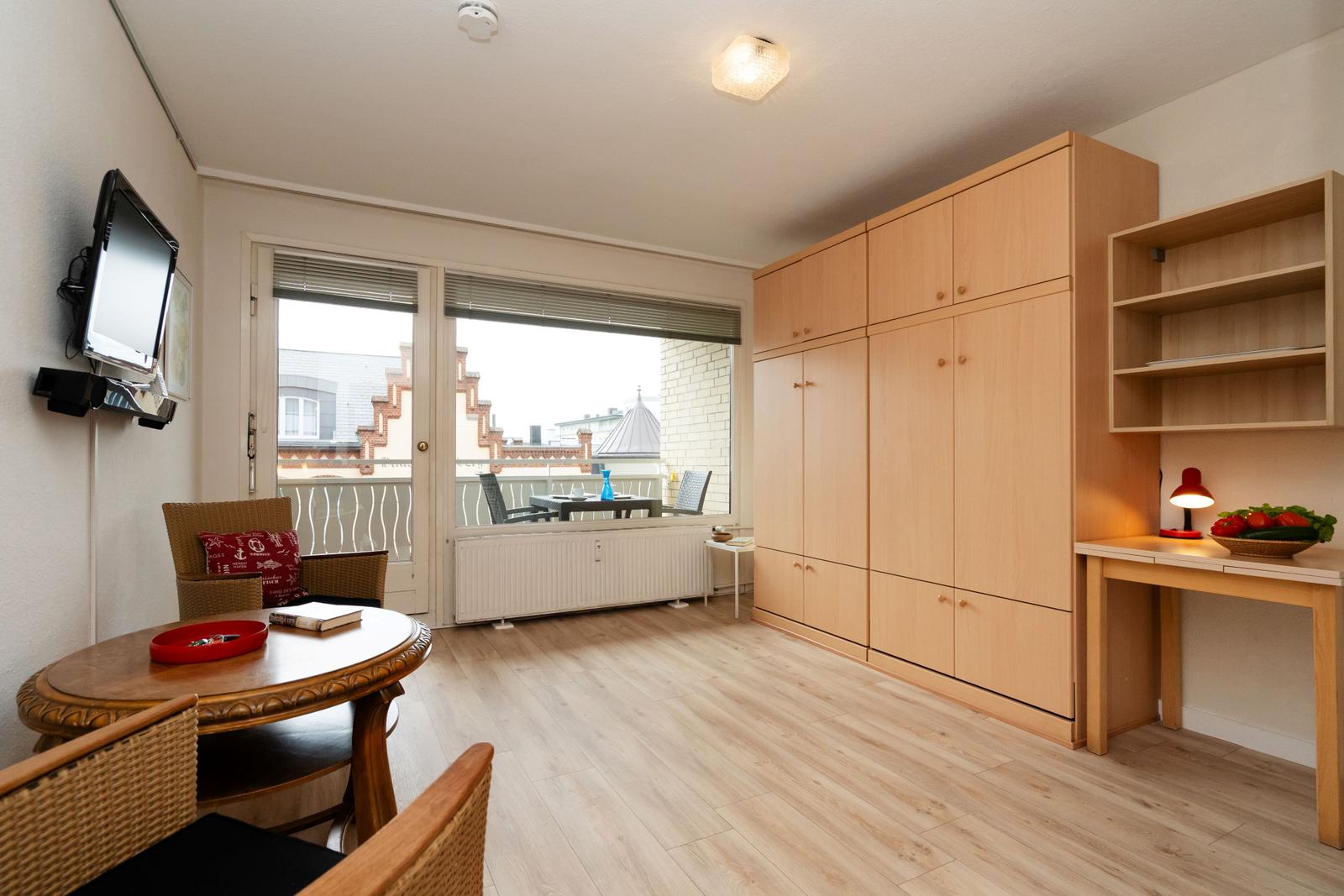 Zimmer mit Holzmöbeln, Balkon und TV. Arbeitsplatz mit Lampe.